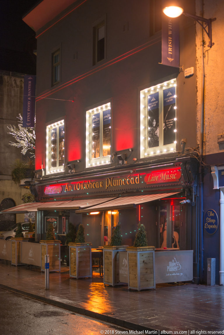 An Brog pub at night with red lighting, awnings, outdoor seating, and reflections on wet pavement.