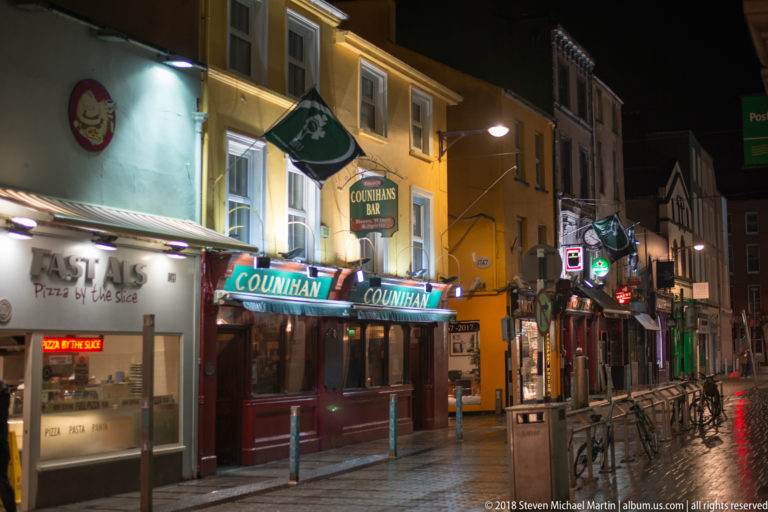 Counihan’s Bar with dark red frontage, illuminated signs, hanging flags, bicycles, and wet street at night.
