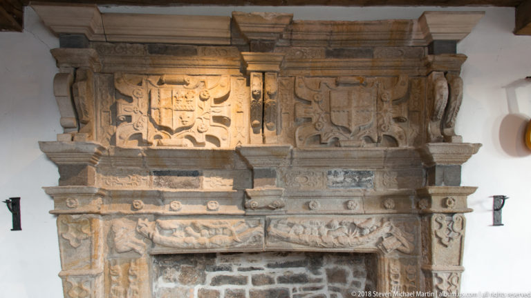 Close view of ornate stone fireplace with heraldic shields, moulded cornices, and carved relief decoration.