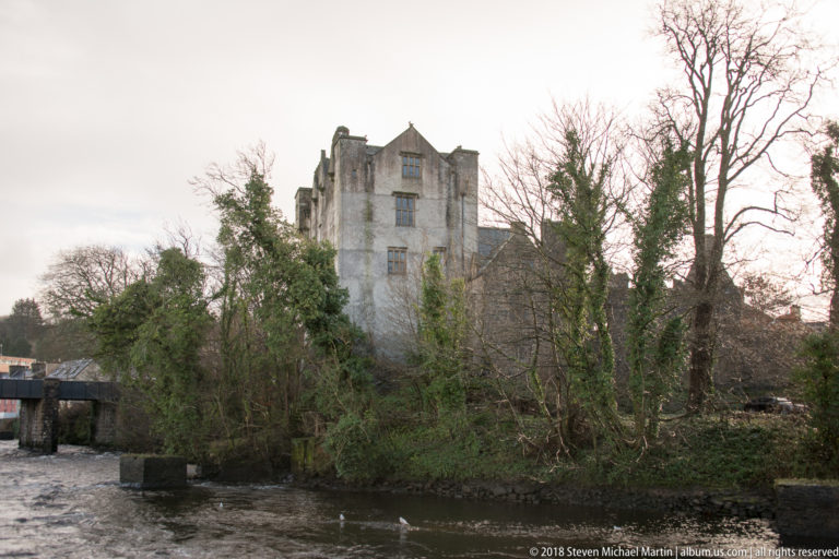 Donegal Castle beyond leafless riverside trees, viewed across flowing water.