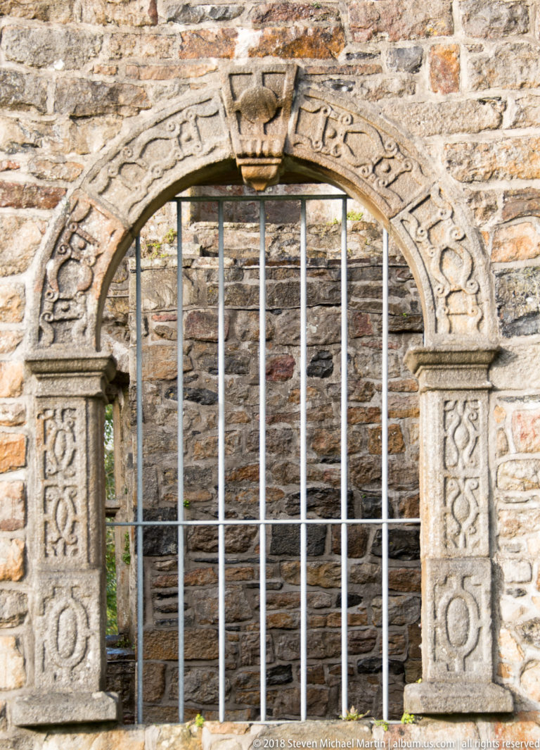 Carved arched stone window surround with geometric decoration, worn lettering, and vertical metal bars.
