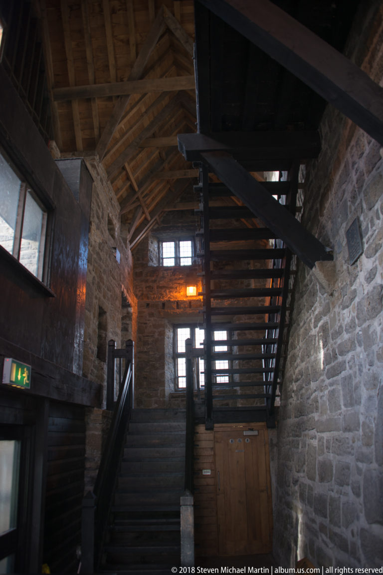 Dark timber staircase rises through a stone-walled interior beneath exposed roof beams.