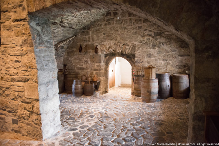Low stone-vaulted room with cobbled floor, wooden barrels, and an arched passage to a bright doorway.