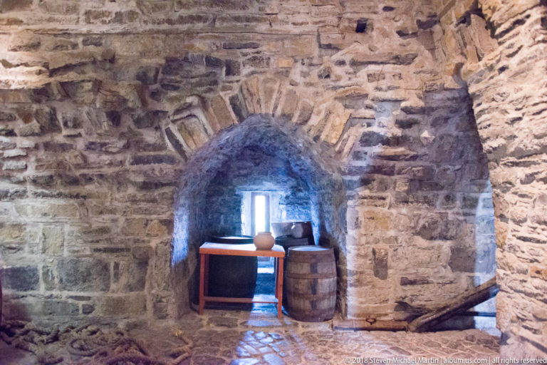 Rough stone chamber with a deep arched recess, small window, display surface, and wooden barrel.