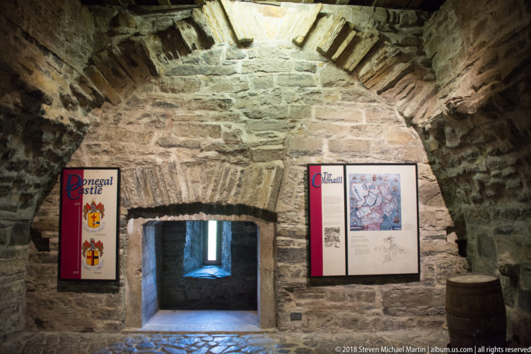 Museum panels and a map flank a low stone doorway in a vaulted castle room.
