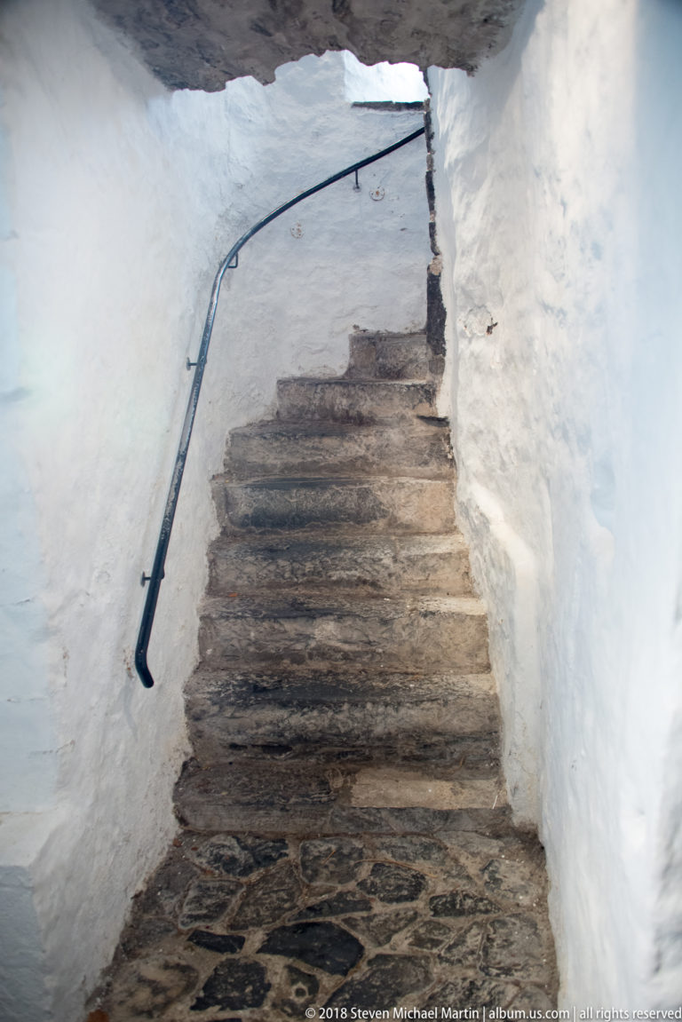Steep worn stone steps rise through a narrow whitewashed passage beside a black iron handrail.
