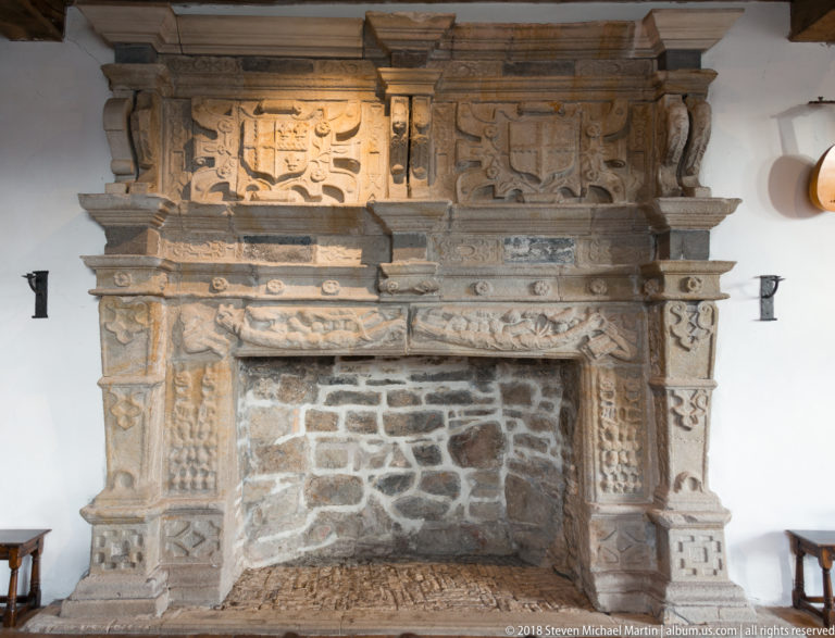 Full frontal view of a monumental carved stone fireplace with shields, relief panels, and a large masonry firebox.