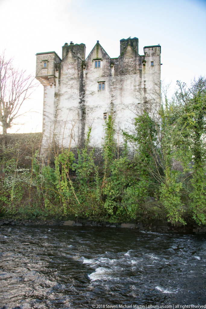 Long pale-stone castle elevation above a dark river, partly screened by dense green vegetation.