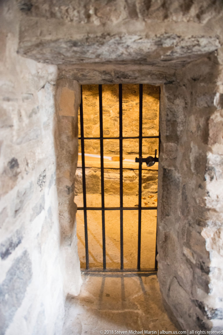 Small rectangular opening with iron bars set deep in a thick rough-stone wall.