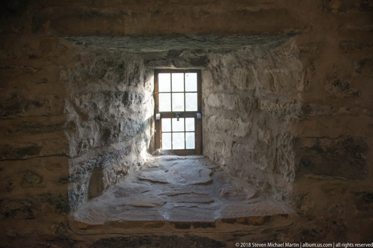 Small multi-pane window at the end of a deep rough-stone reveal with a broad sill.