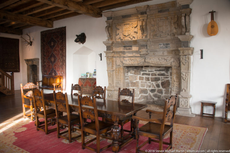 Long wooden dining table and carved chairs face an ornate stone fireplace in a restored castle hall.