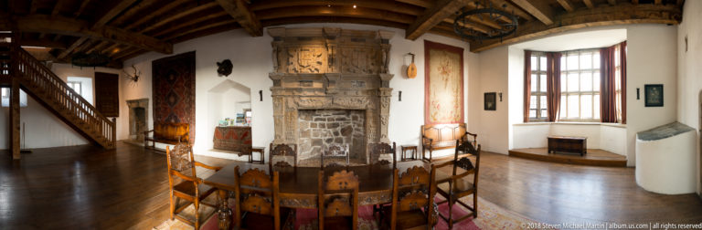 Wide restored hall with timber ceiling, carved stone fireplace, long table, chairs, tapestries, and tall windows.