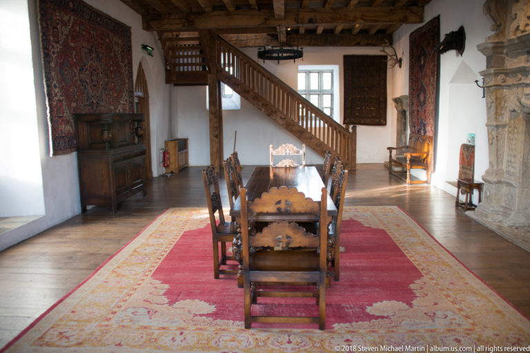 Long table and carved chairs on a red carpet, with staircase, tapestries, and fireplace edge beyond.