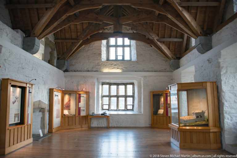 High stone-walled gallery with exposed roof timbers, large windows, and museum display cases around an open floor.