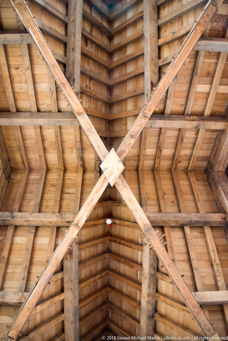 Upward view of a timber roof with crossing diagonal braces, rafters, beams, and closely spaced wooden boards.
