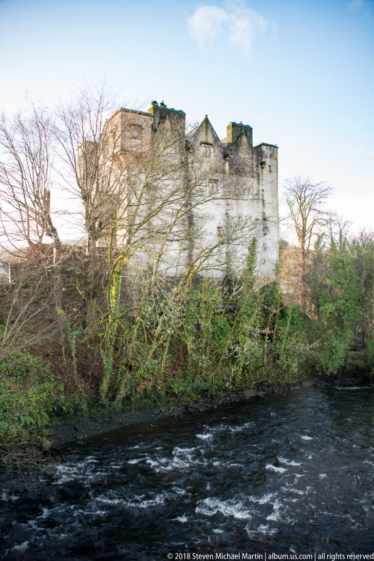 Oblique view of Donegal Castle behind riverside trees, with rippling water in the foreground.