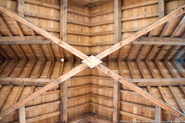 Close upward view of diagonal timber braces crossing beneath boarded roof framing with rafters and horizontal beams.