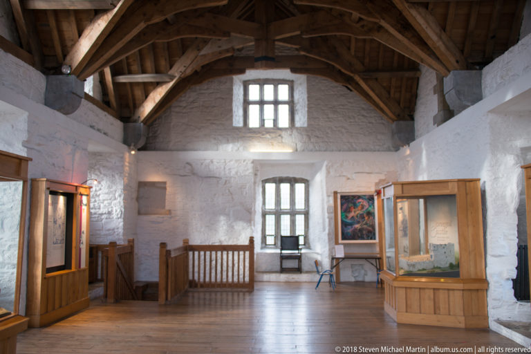 Restored castle room with white masonry walls, exposed timber roof, plank floor, deep windows, and museum display cases.