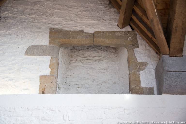 Close view of a rectangular stone fireplace opening in a white masonry wall beneath heavy timber roof members.