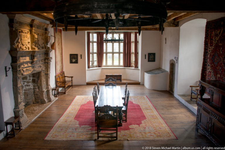 Castle room with long wooden dining table, red patterned carpet, carved stone fireplace, ceiling beams, windows, and historic furnishings.
