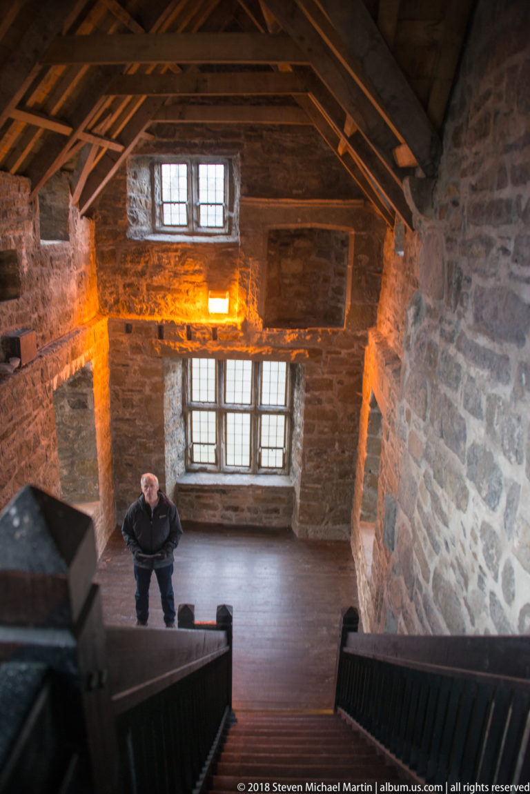 Downward view from a timber staircase into a tall stone hall with two multi-pane windows and a person standing below.