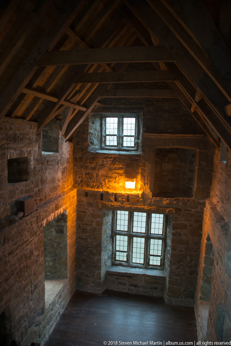 Empty stone castle chamber with dark timber roof framing and two vertically aligned multi-pane windows in the far wall.