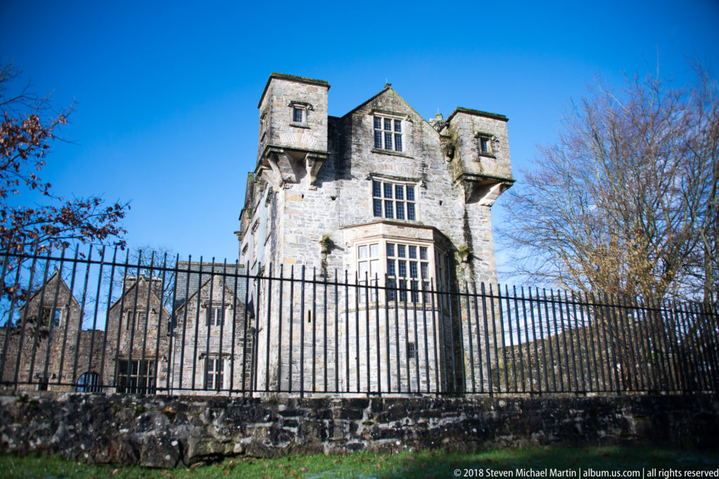 Tall stone corner of Donegal Castle with projecting upper rooms behind a black iron fence.