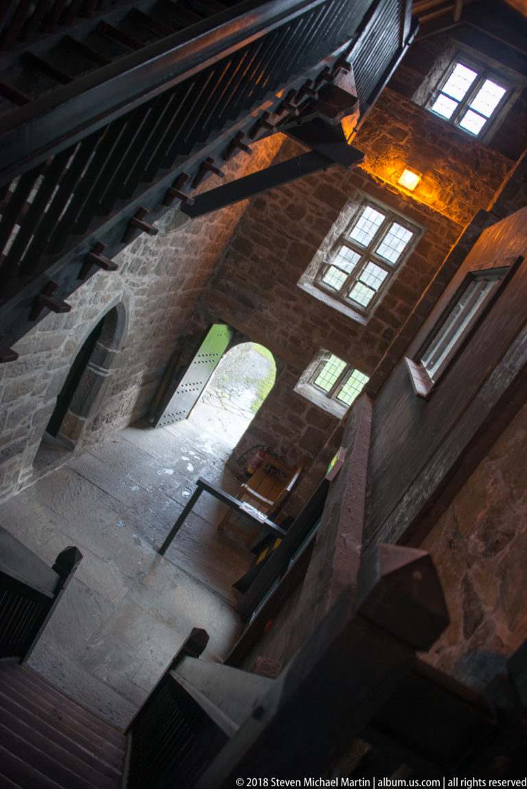 Tilted interior view of a steep timber staircase, stone walls, recessed windows, and an arched doorway opening outdoors.
