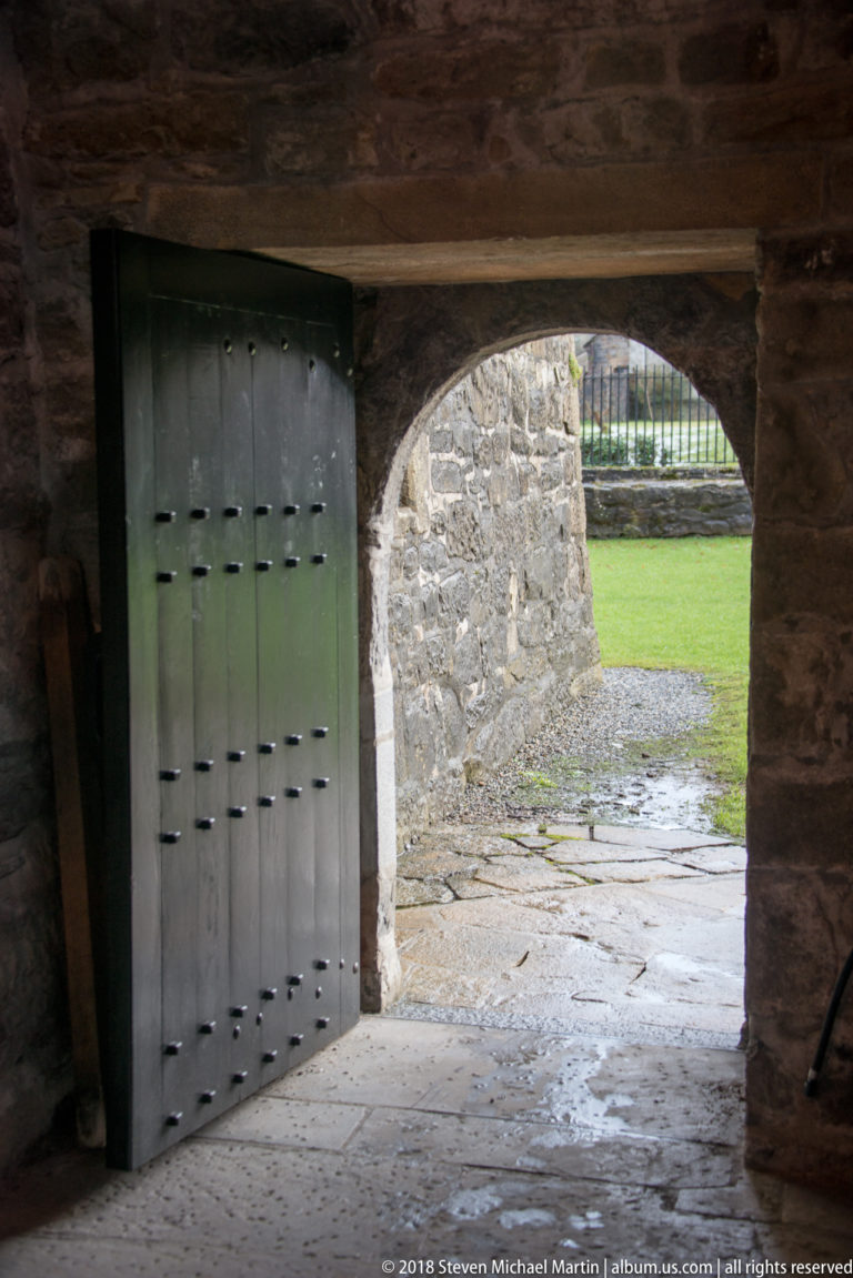 Partly open studded timber door inside a stone passage, with an arched opening and green lawn visible beyond.