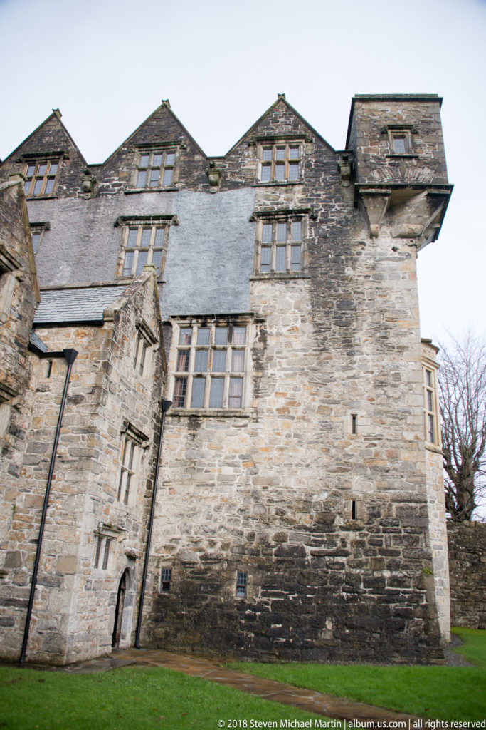 Stone castle elevation with multi-light windows, steep rooflines, projecting bays, and weathered masonry above a green lawn.