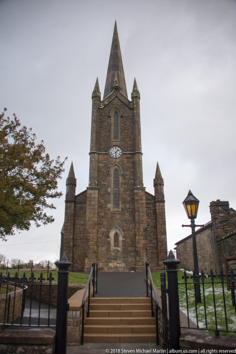 Front view of a stone Gothic Revival church with pointed tower, clock face, pinnacles, steps, and iron railings.