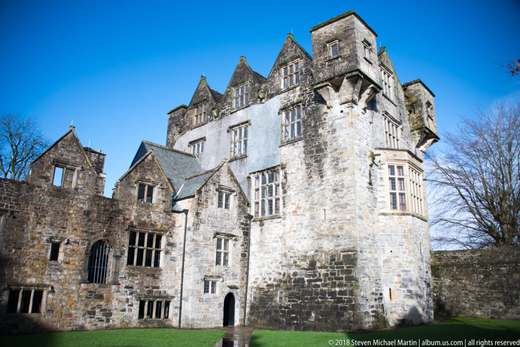 Sunlit stone tower with mullioned windows beside lower roofed and partly open castle ranges.