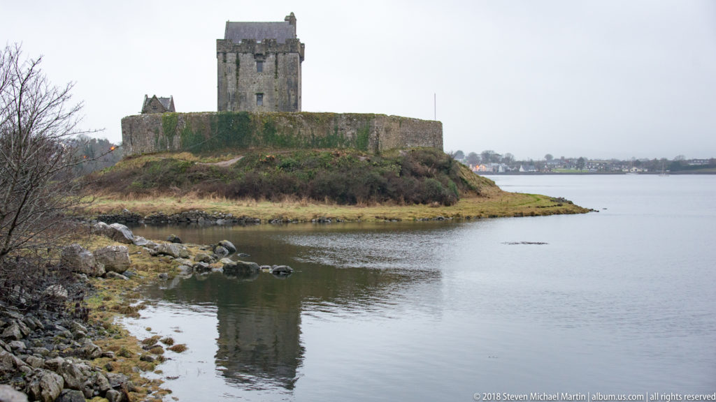 Dunguaire Castle on a low peninsula beside calm water, with tower house, curtain wall, rocky shore, and soft reflection.