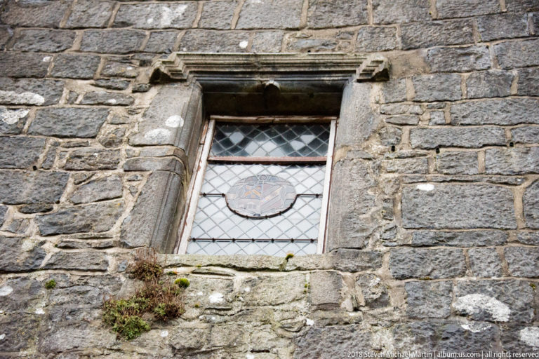 Leaded window with diamond panes and a small central decorative panel set in the rough stone wall of Dunguaire Castle.
