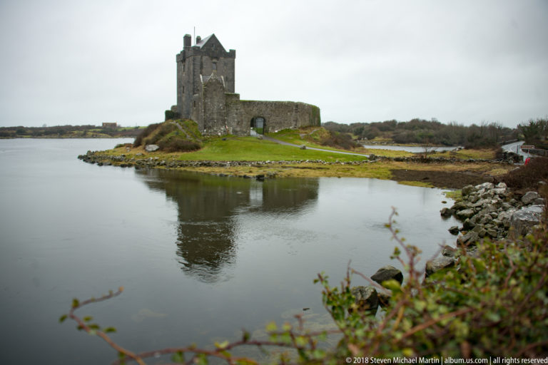 Dunguaire Castle stands across still water with a soft reflection, framed by shoreline rocks and low vegetation.