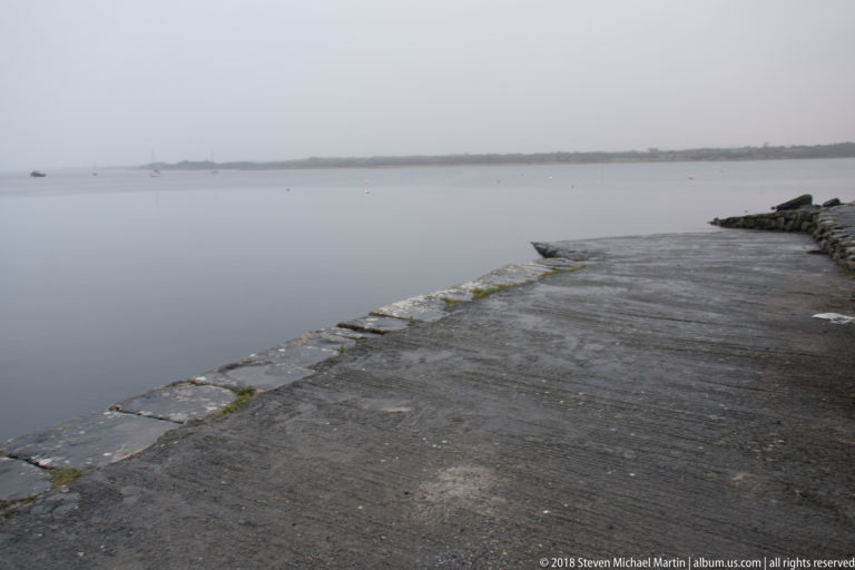 Concrete harbor slipway bordered by flat stones descends into calm grey water under an overcast sky.
