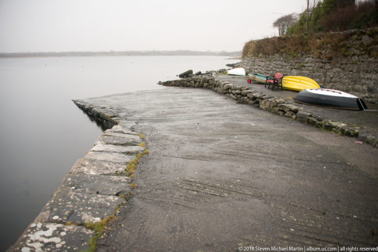 Harbor slipway beside a stone quay with bright kayaks and a small dinghy pulled up along the water’s edge.