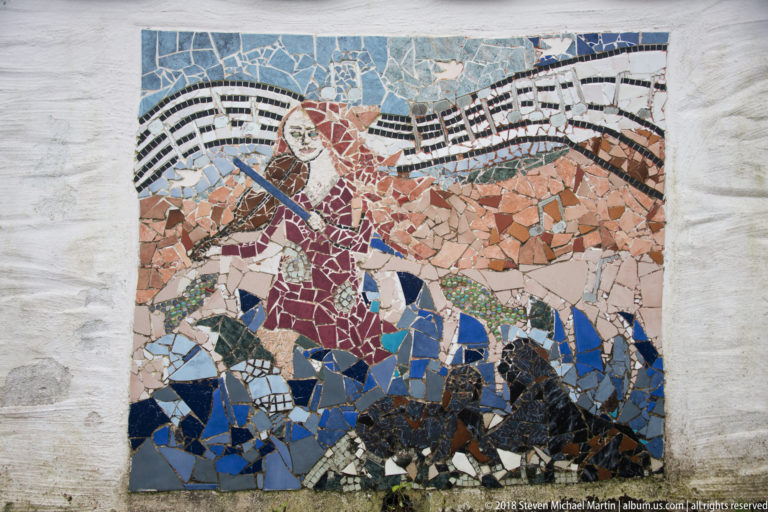 Colorful broken-tile mosaic in a white wall, showing a stylized human figure with a large brown animal form.