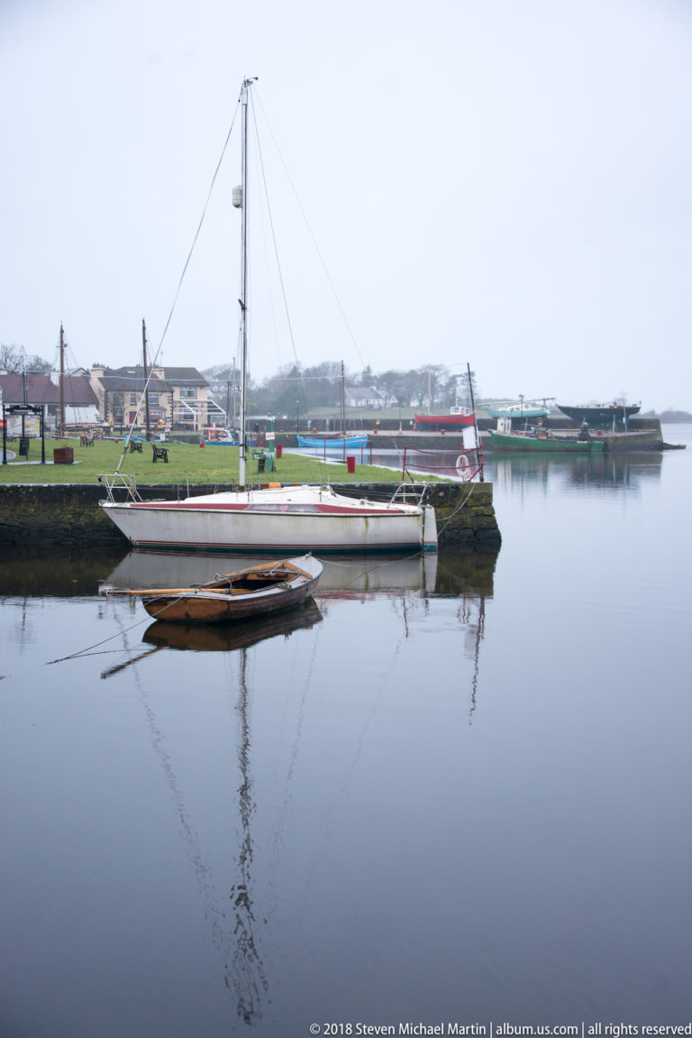 White sailboat and smaller wooden boat moored in calm harbor water, with reflections and low waterfront buildings behind.