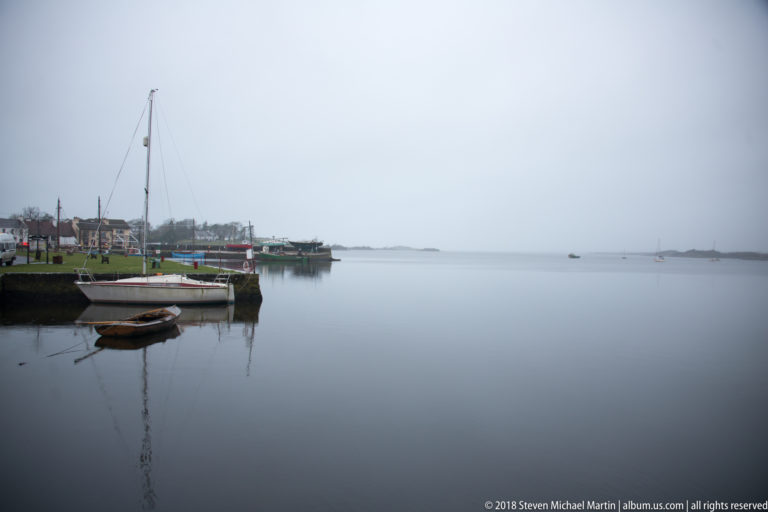 Wide view across calm Kinvara harbor with moored sailboats, quay buildings at left, and open grey water extending into haze.