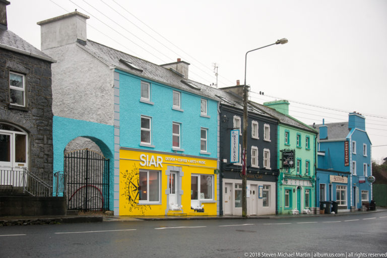 Colorful two-story buildings and storefronts line a wet street in Kinvara, including turquoise, yellow, grey, and green façades.
