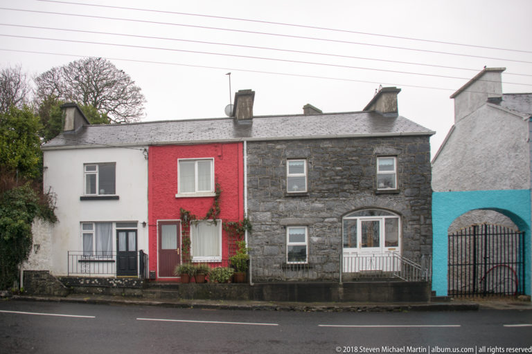 Row of Kinvara houses with white, red, grey stone, and turquoise façades beneath slate roofs and chimneys.
