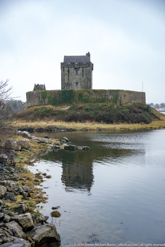 Distant side view of Dunguaire Castle across calm water, with the tower house behind a long stone enclosure wall.