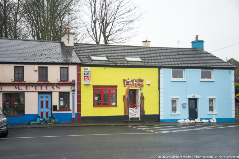 Colorful Kinvara storefronts include a cream-and-blue frontage, bright yellow café, and pale blue adjoining building under slate roofs.
