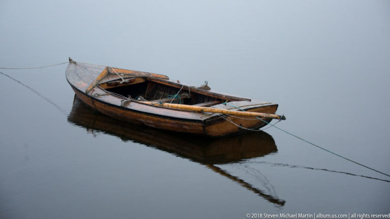 Weathered open wooden boat with visible ribs and benches floats on still water, tethered by mooring lines and reflected below.