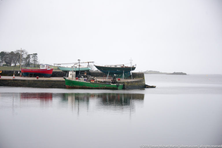 Green working boat beside a quay with several larger boats hauled out on supports, reflected in calm harbor water.