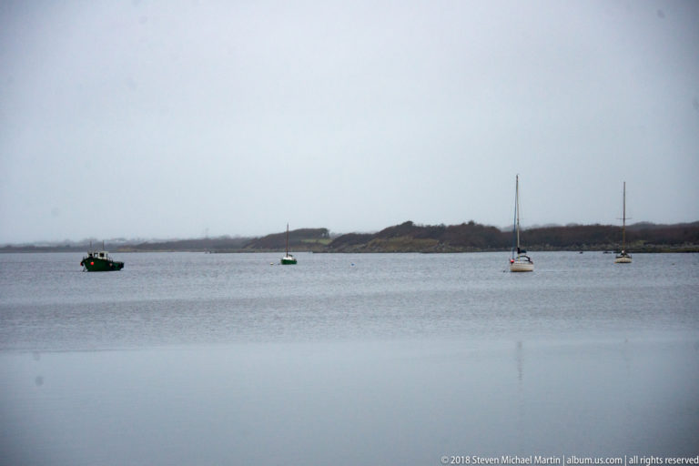 Small motorboat and several moored sailboats are spaced across calm water before a low dark shoreline.