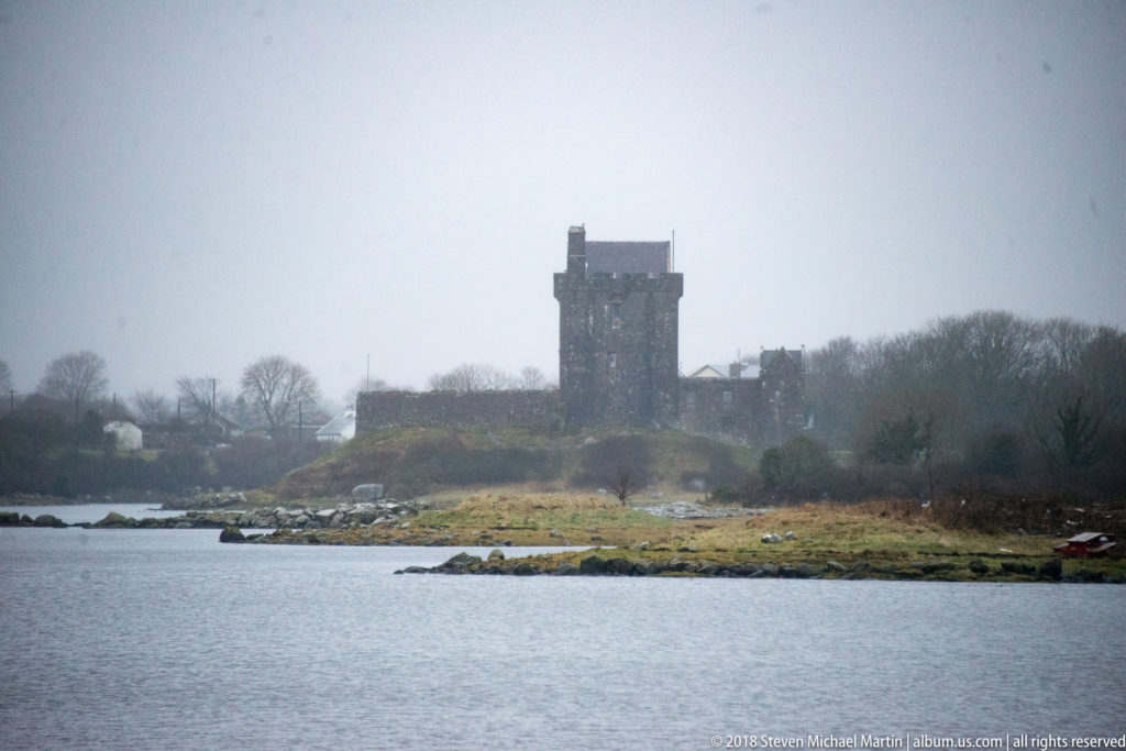 Dunguaire Castle seen through mist across grey water, with its dark tower house rising above the low shoreline and trees.