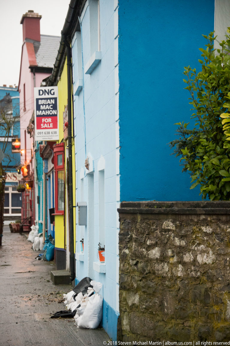 Narrow Kinvara lane between brightly painted blue, yellow, and pink buildings, with a projecting accommodation sign overhead.