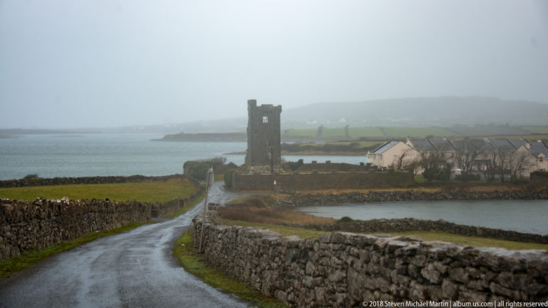 Ruined square stone tower beside a narrow wet road and houses across an inlet, with drystone walls and misty hills beyond.